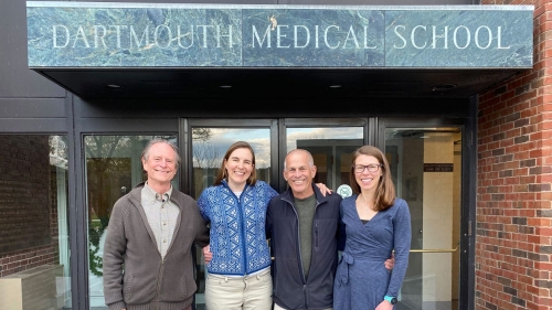 Four people stand with their arms around each other in front of the Dartmouth Medical School entrance.