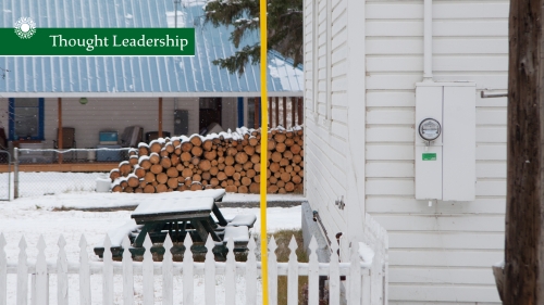 An electricity meter on a white house with firewood piled outside and dusting of snow.