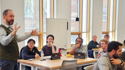 Students sit at tables and listen to an instructor who speaks and gestures with his hands.