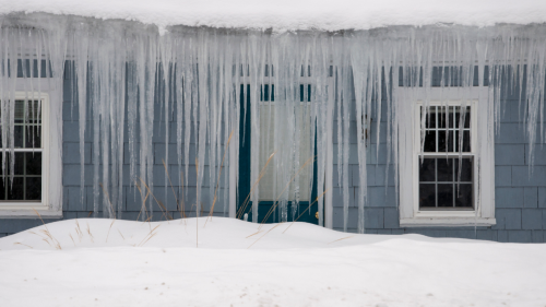 a stock photo of an old blue farmstyle house in the snow with massive stalagmite like icicles hanging from the roof