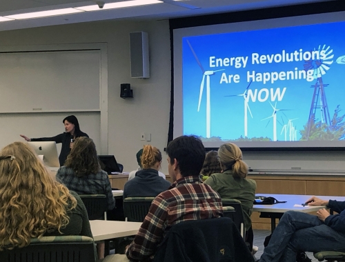 Image of Elizabeth Wilson at head of classroom a screen with a windmill on it that says Energy Revolutions are Happening Now class full of students