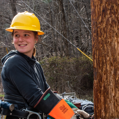 A young woman wearing a hard hat and smiling as she scales a utility pole