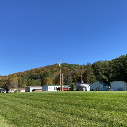 Green grass in the foreground, a row of mobile homes in the middle ground, and a small, fall foliage covered hill in the background with a blue sky