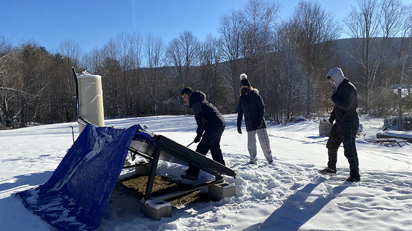 Three students set up a solar panel and water heater in a snowy field