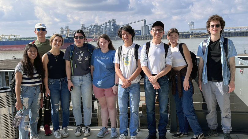 A group of students posing in the gulf coast with an oil tanker behind it