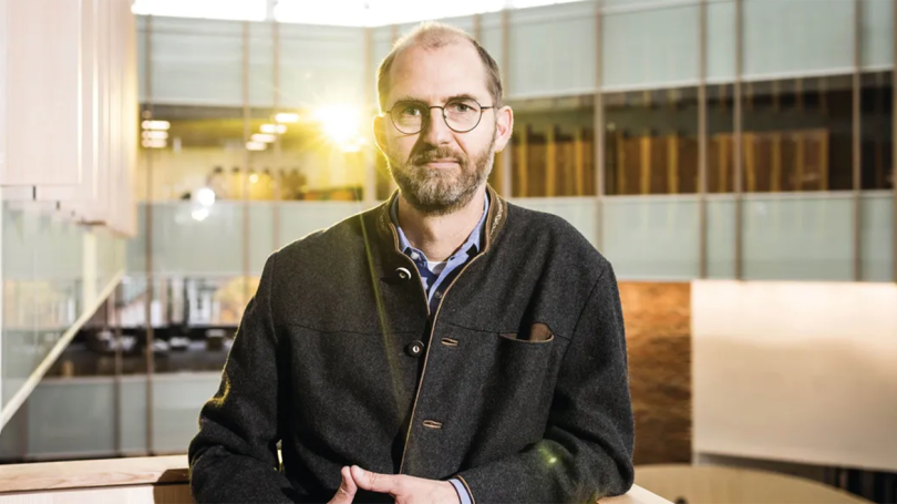 Klaus Keller standing against a low wall with Irving Institute atrium in background