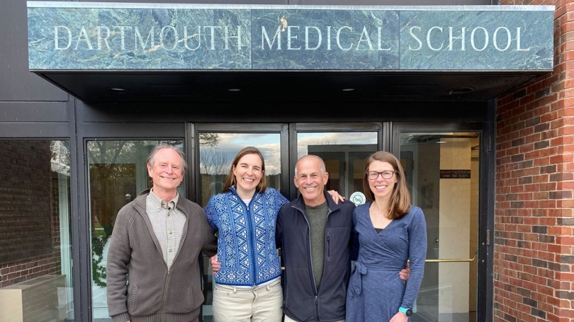 Four people stand with their arms around each other in front of the Dartmouth Medical School entrance.