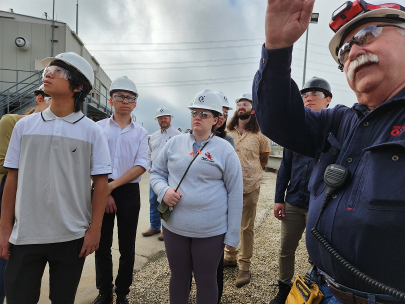 Students in hard hats and safety goggles listening to a man talking at an energy facility