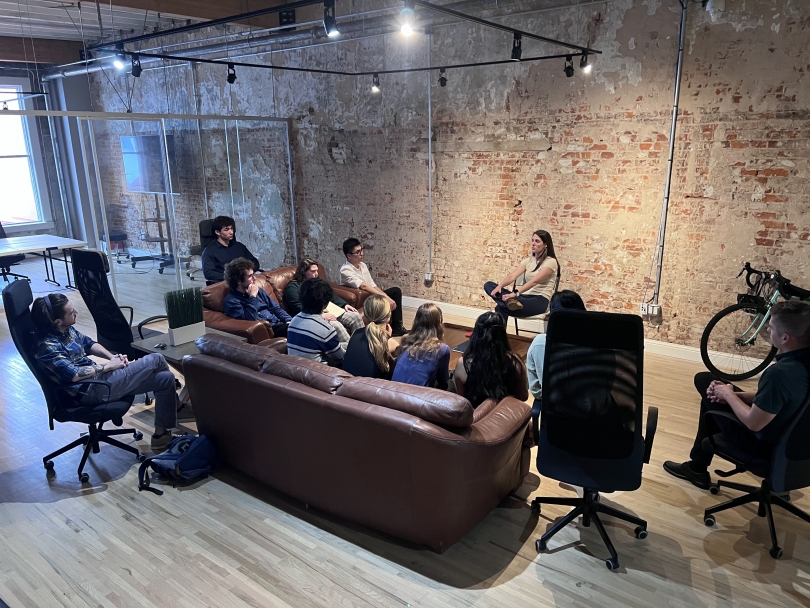 Students sitting on a couch and chairs in a brick and glass walled space listening to a woman talking at the front of the room 
