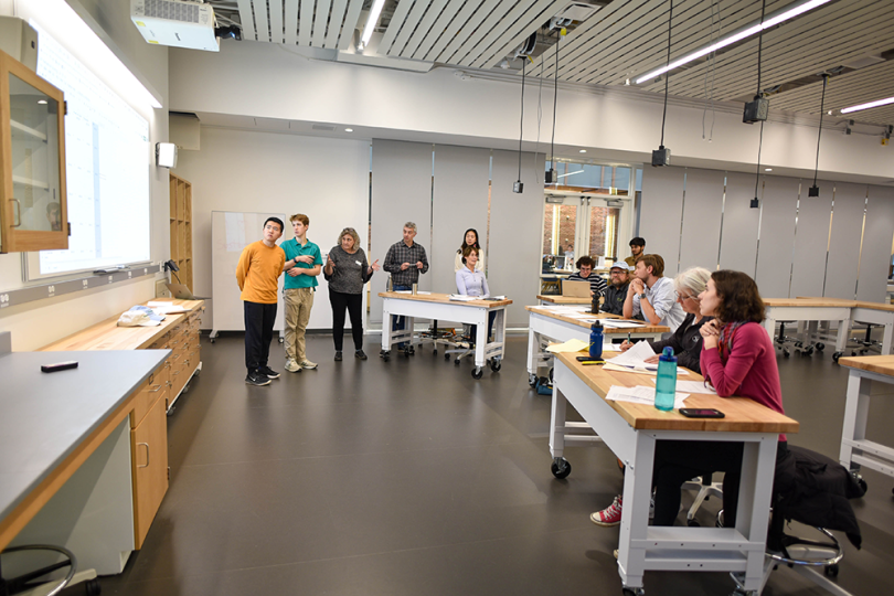 People sitting at a table listening to students standing in front of a lab
