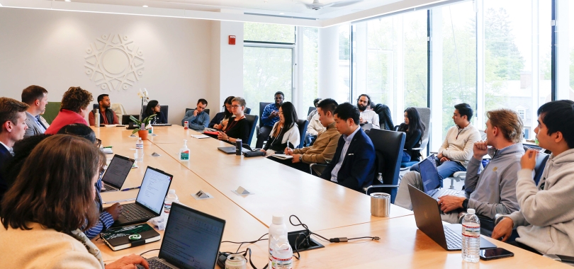 Students and industry professionals sit at a large table in a light-filled board room.