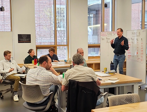 Students sit at tables and listen to an instructor who speaks in front of whiteboards.