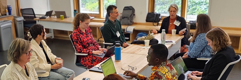 A group of people sit at a table with laptops, talking with each other.