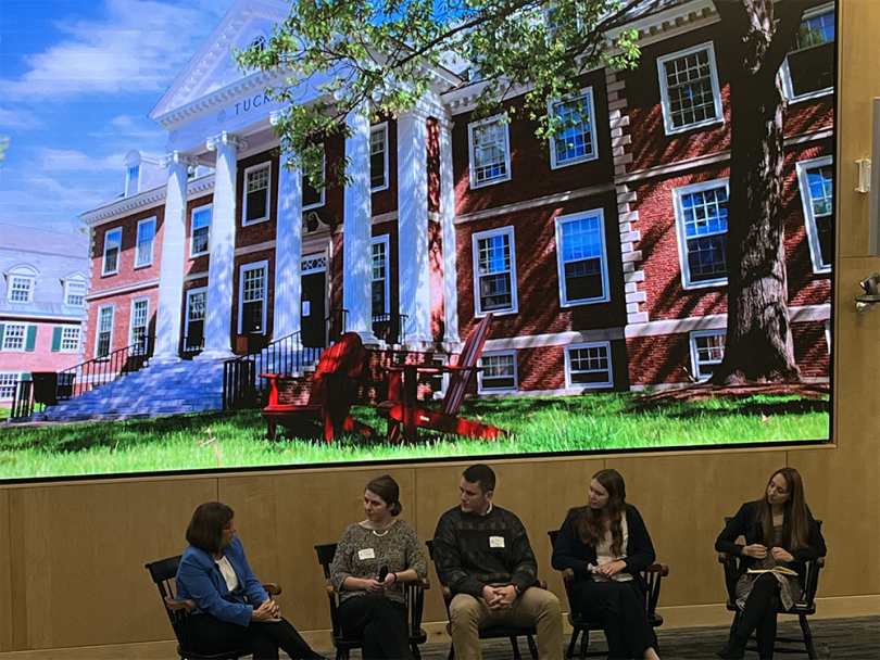 A group of people sitting on a stage beneath a large photo of the Tuck School of Business