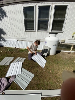 A person in front of a house near a propane tank with several pieces of corrugated metal on the ground surrounding them; the person is cutting a piece of corrugated metal