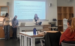 Three students deliver a presentation in front of a projector screen with the word 'Goals' and several bullet points of text.