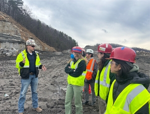 A worker a students stand in a gravel yard wearing safety vests and hardhats.